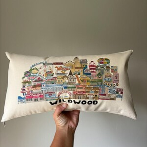 K&ouml;nnte beinhalten: Ein wei&szlig;es Kissen mit einer bunten Illustration der Wildwood Promenade. Die Illustration zeigt Geb&auml;ude, Fahrgesch&auml;fte und Schilder mit Texten wie "Wawa", "Morey's Piers" und "Diner". Das Kissen hat das Wort "WILDWOOD" in Schwarz unten aufgedruckt.