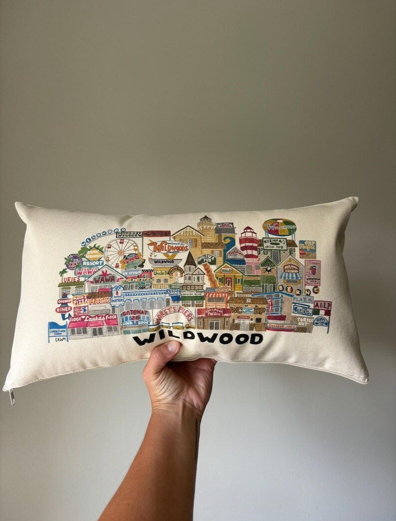 K&ouml;nnte beinhalten: Ein wei&szlig;es Kissen mit einer bunten Illustration der Wildwood Promenade. Die Illustration zeigt Geb&auml;ude, Fahrgesch&auml;fte und Schilder mit Texten wie "Wawa", "Morey's Piers" und "Diner". Das Kissen hat das Wort "WILDWOOD" in Schwarz unten aufgedruckt.