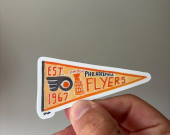 May include: A pennant-shaped sticker featuring the Philadelphia Flyers logo. The sticker is orange and white with the text "EST. 1967" and "PHILADELPHIA FLYERS". The logo includes a stylized "P" and a hockey puck.