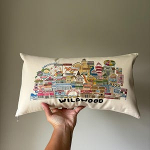 K&ouml;nnte beinhalten: Ein wei&szlig;es Kissen mit einer bunten Illustration der Wildwood Promenade. Die Illustration zeigt Geb&auml;ude, Gesch&auml;fte, Fahrgesch&auml;fte und den Text "Wildwood".