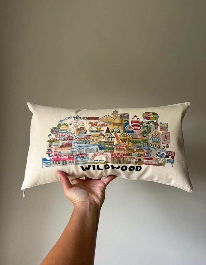 K&ouml;nnte beinhalten: Ein wei&szlig;es Kissen mit einer bunten Illustration der Wildwood Promenade. Die Illustration zeigt Geb&auml;ude, Gesch&auml;fte, Fahrgesch&auml;fte und den Text "Wildwood".