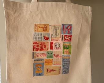 May include: A beige canvas tote bag with a colorful collage of baseball-themed illustrations. The collage includes images of a pretzel, baseball caps, and the text "Phillies" and "Ring the Bell". The bag has two beige handles.