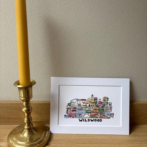 May include: A white framed print with a colorful illustration of the Wildwood boardwalk. The illustration includes a Ferris wheel, a lighthouse, and other buildings. The text "WILDWOOD" is printed below the illustration.