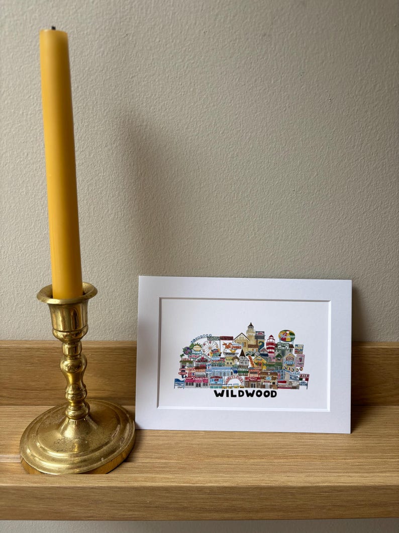 May include: A white framed print with a colorful illustration of the Wildwood boardwalk. The illustration includes a Ferris wheel, a lighthouse, and other buildings. The text "WILDWOOD" is printed below the illustration.