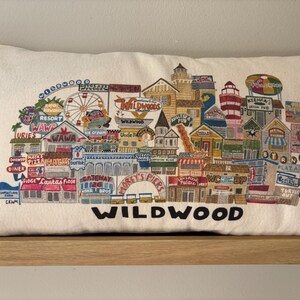K&ouml;nnte beinhalten: Ein wei&szlig;es Kissen mit einem bunten gestickten Design der Wildwood Promenade. Das Design umfasst verschiedene Gesch&auml;fte, Restaurants und Attraktionen, darunter Wawa, Doo Wop Diner und Morey's Piers. Der Text "WILDWOOD" ist in Schwarz am unteren Rand des Kissens gestickt.