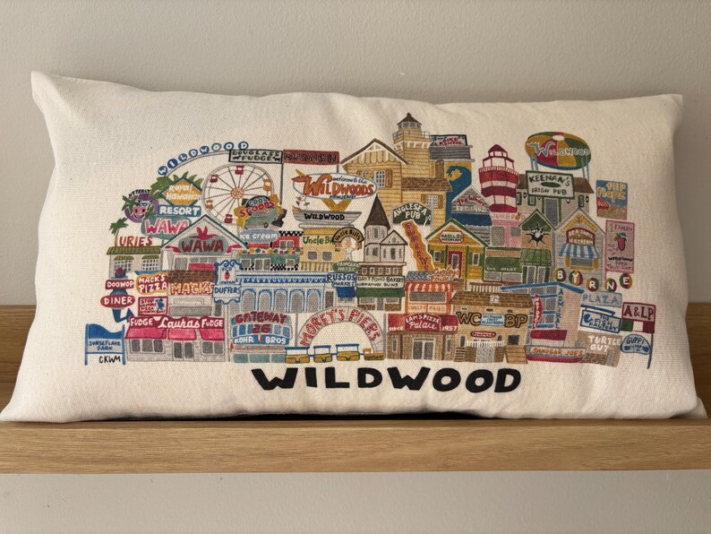 K&ouml;nnte beinhalten: Ein wei&szlig;es Kissen mit einem bunten gestickten Design der Wildwood Promenade. Das Design umfasst verschiedene Gesch&auml;fte, Restaurants und Attraktionen, darunter Wawa, Doo Wop Diner und Morey's Piers. Der Text "WILDWOOD" ist in Schwarz am unteren Rand des Kissens gestickt.