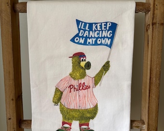 May include: White kitchen towel with a green, furry cartoon bear wearing a red and white baseball jersey with the word "Phillies" on it. The bear is holding a blue flag that says "I'll keep dancing on my own."