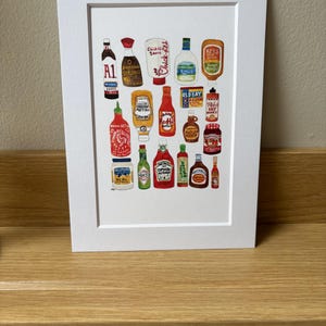 Op de afbeelding: Een aquarelillustratie van een verzameling verschillende condimentenflessen, waaronder ketchup, mosterd, hete saus, sojasaus en mayonaise. De flessen zijn gerangschikt in een rasterpatroon op een witte achtergrond.