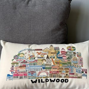K&ouml;nnte beinhalten: Ein wei&szlig;es Kissen mit einer bunten Illustration der Wildwood Promenade, die verschiedene Gesch&auml;fte, Restaurants und Attraktionen zeigt. Der Text "WILDWOOD" ist unten in Schwarz gedruckt.