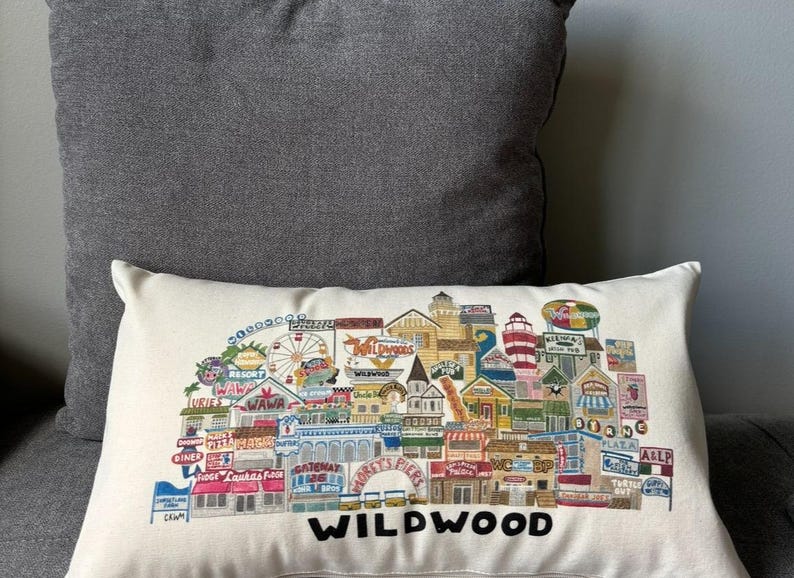 K&ouml;nnte beinhalten: Ein wei&szlig;es Kissen mit einer bunten Illustration der Wildwood Promenade, die verschiedene Gesch&auml;fte, Restaurants und Attraktionen zeigt. Der Text "WILDWOOD" ist unten in Schwarz gedruckt.
