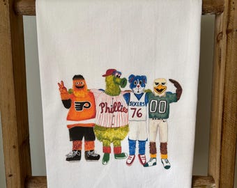 May include: A white kitchen towel with a colorful illustration of four mascots. The mascots are wearing jerseys with the words "Phillies", "Sixers", and "00".