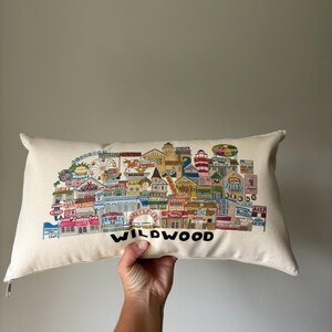 K&ouml;nnte beinhalten: Ein wei&szlig;es Kissen mit einer bunten Illustration der Wildwood Promenade. Die Illustration zeigt Geb&auml;ude, Gesch&auml;fte, Fahrgesch&auml;fte und den Text "Wildwood".