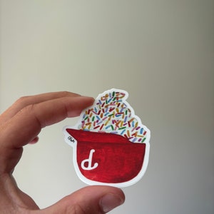 Op de afbeelding: Een rode en witte sticker van een rode beker gevuld met kleurrijke sprinkels. De beker heeft een witte letter "D" aan de voorkant.