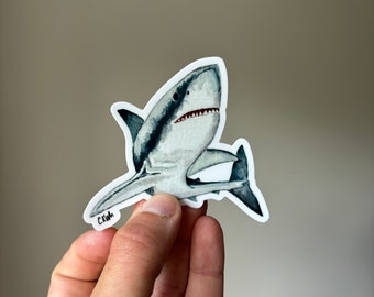 May include: A white shark sticker with a watercolor design. The shark is facing forward with its mouth open and teeth visible. The sticker has a white border and the artist's signature "CKWH" in the bottom left corner.
