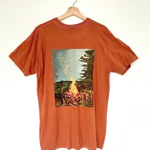 May include: Burnt orange t-shirt with a rectangular graphic of a campfire scene. The graphic features a bonfire with flames, a starry sky, and the text "I Consider That Joy". The shirt is hanging on a wooden hanger.