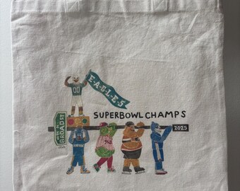 May include: Cream-colored tote bag with beige handles featuring a colorful illustration. The illustration includes the text "E-A-G-L-E-S" on a banner, "SUPERBOWL CHAMPS" and the year "2025". The bag has a rectangular shape.