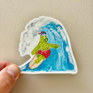 May include: A sticker featuring a green character surfing a blue wave. The character wears a red cap and red swim trunks. The surfboard is tan. The sticker is die-cut with a white border.