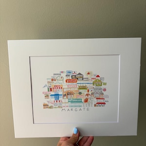 May include: A white matted art print featuring a colorful, detailed illustration of various storefronts and landmarks in Margate, including an elephant figure, with the word MARGATE printed in black capital letters at the bottom center of the artwork.