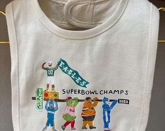 May include: Cream-colored baby bib with a colorful graphic. The graphic features cartoon characters carrying a banner that reads "E-A-G-L-E-5" and the text "SUPERBOWL CHAMPS 2025". The bib has a snap closure at the neck and is hanging on a gold hanger.
