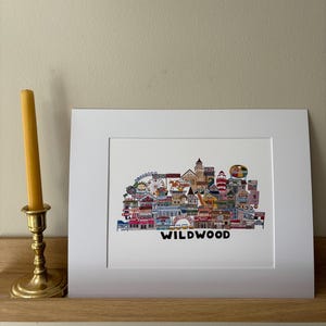 May include: A white framed print of a colorful illustration of the Wildwood boardwalk in New Jersey. The illustration is made up of various buildings, shops, and attractions, including a Ferris wheel, a roller coaster, and a lighthouse. The text "WILDWOOD" is printed below the illustration.