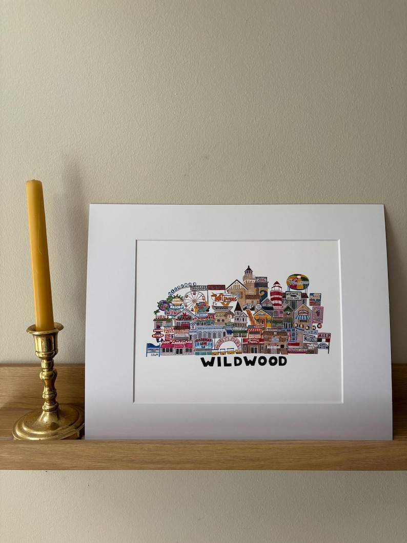May include: A white framed print of a colorful illustration of the Wildwood boardwalk in New Jersey. The illustration is made up of various buildings, shops, and attractions, including a Ferris wheel, a roller coaster, and a lighthouse. The text "WILDWOOD" is printed below the illustration.