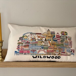 K&ouml;nnte beinhalten: Ein wei&szlig;es Kissen mit einem bunten gestickten Design der Wildwood Promenade. Das Design beinhaltet Geb&auml;ude, ein Riesenrad und den Text "Wildwood".
