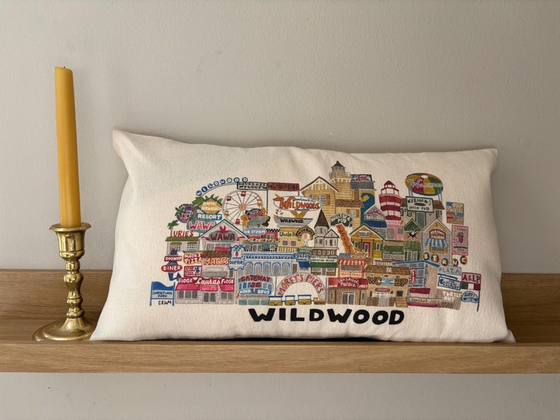 K&ouml;nnte beinhalten: Ein wei&szlig;es Kissen mit einem bunten gestickten Design der Wildwood Promenade. Das Design beinhaltet Geb&auml;ude, ein Riesenrad und den Text "Wildwood".