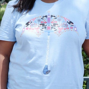 May include: A woman wearing a light blue t-shirt with a colorful umbrella design. The umbrella is made up of various items, including shoes, hats, and purses. The woman is smiling and wearing sunglasses.