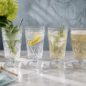 May include: Four clear glass goblets with a diamond-patterned texture on the upper bowl and a square base. Each glass contains a different beverage with garnishes including fresh mint, a lemon twist, a rosemary sprig, and a single raspberry.
