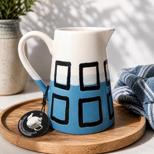 May include: A ceramic pitcher featuring a white upper section and a blue lower section with a hand-painted black rectangular grid pattern. A round black tag with white text reading The Old Pottery Company Designed In England is attached.