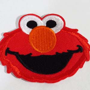 Op de afbeelding: Een rood geborduurd embleem met het gezicht van een stripfiguur met witte ogen, zwarte pupillen, een oranje neus en een grote zwarte lachende mond. De patch heeft een getextureerd uiterlijk en is bedoeld voor kleding of accessoires.