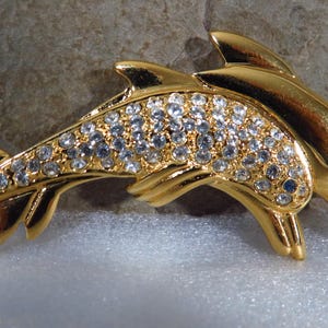 May include: Gold-toned dolphin brooch with clear crystal accents and blue eyes. The brooch features two dolphins, one slightly behind the other, with their bodies curved. A classic jewelry piece.