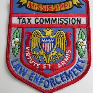 Puede incluir: Parche bordado con las palabras "MISSISSIPPI TAX COMMISSION LAW ENFORCEMENT". El parche tiene un fondo azul, un borde rojo y un emblema central de un águila dorada. También incluye las palabras "Virtute et Armis".