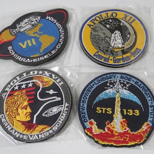 Op de afbeelding: Vier geborduurde NASA-missiepatches in doorzichtige plastic hoesjes. Linksboven: Apollo VII met aarde en ruimtevaartuig. Rechtsboven: Apollo XII met een schip. Linksonder: Apollo XVII met een profiel van Apollo. Rechtsonder: STS-133 met een spaceshuttle.