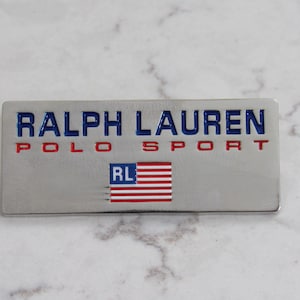 Op de afbeelding: Een rechthoekig zilverkleurig metalen embleem met de woorden "RALPH LAUREN POLO SPORT" in blauwe en rode letters. Onder de tekst staat een kleine Amerikaanse vlag met de letters "RL".