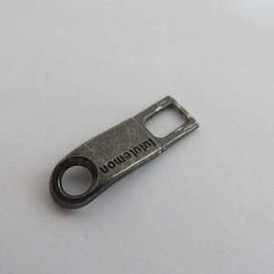 Puede incluir: Un pequeño tirador de cremallera metálico gris con la palabra "lululemon" impresa. Tiene una abertura rectangular en un extremo y una abertura circular en el otro. La superficie tiene un aspecto ligeramente desgastado.