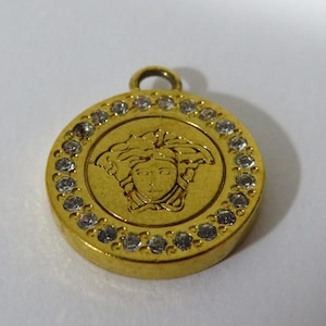 Puede incluir: Colgante dorado con diseño circular. El colgante presenta una imagen central en relieve de una cara, rodeada por un anillo de pequeñas piedras preciosas transparentes. El colgante tiene un lazo en la parte superior.