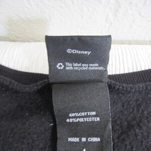 Peut inclure: Gros plan sur une &eacute;tiquette de v&ecirc;tement noire avec du texte blanc. L'&eacute;tiquette indique "&copy;Disney", "This label was made with recycled materials," et "60%COTTON 40%POLYESTER MADE IN CHINA 1."
