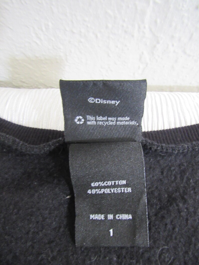 Peut inclure: Gros plan sur une &eacute;tiquette de v&ecirc;tement noire avec du texte blanc. L'&eacute;tiquette indique "&copy;Disney", "This label was made with recycled materials," et "60%COTTON 40%POLYESTER MADE IN CHINA 1."