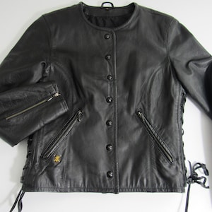 Może przedstawiać: Czarna skórzana kurtka z okrągłym kołnierzem, zapięciem na guziki i kieszeniami na zamek błyskawiczny. Kurtka posiada sznurowanie po bokach i małą złotą odznakę. Rękawy mają zamki przy mankietach.