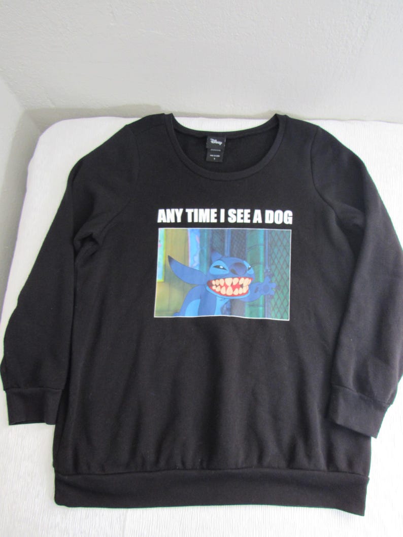 Peut inclure: Sweat-shirt noir &agrave; manches longues avec un dessin du personnage Disney Stitch et le texte "ANY TIME I SEE A DOG". L'image montre Stitch avec un grand sourire. Le sweat-shirt a un col rond et des poignets c&ocirc;tel&eacute;s.