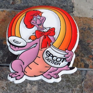Puede incluir: Un pin colorido de estilo caricatura que presenta un dragón púrpura con cabello rojo y una pajarita. El dragón sostiene una olla con la etiqueta "EPCOT CENTER" y un plato pequeño, con un arcoíris arqueándose detrás. El pin está sobre un fondo de piedra texturizada.