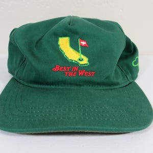 Puede incluir: Gorra de béisbol verde con visera curva. La gorra presenta un diseño bordado de un campo de golf en la parte delantera, con las palabras "Best in the West" en rojo. Una pequeña nube verde está bordada en el lateral.