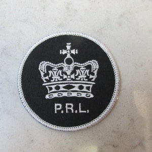 May include: A round, black patch with a white embroidered crown design and the letters "P.R.L." The patch has a white stitched border. The crown is detailed with ornate elements.
