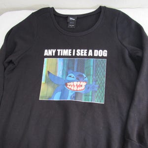 Peut inclure: Sweat-shirt noir &agrave; manches longues avec un motif de Stitch de Lilo & Stitch de Disney. L'image montre Stitch avec un large sourire et le texte "ANY TIME I SEE A DOG" est imprim&eacute; au-dessus du motif. Le sweat-shirt a un col rond.