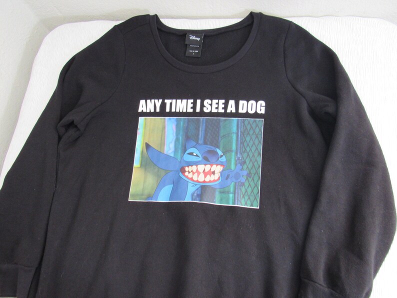 Peut inclure: Sweat-shirt noir &agrave; manches longues avec un motif de Stitch de Lilo & Stitch de Disney. L'image montre Stitch avec un large sourire et le texte "ANY TIME I SEE A DOG" est imprim&eacute; au-dessus du motif. Le sweat-shirt a un col rond.