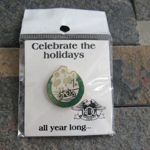 May include: A white card with green text that reads "Celebrate the holidays all year long..." and a gold pin with a green and white design. The pin features a motorcycle and the text "Harley Owners Group 1983".