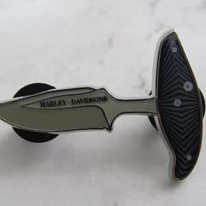 May include: A silver and black Harley-Davidson knife pin with a black and white patterned handle. The knife has a silver blade with the words "Harley-Davidson" engraved on it.