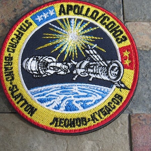 May include: A yellow and red embroidered patch with the text "APOLLO-SOYUZ" and "LEONOV-K.KONRADS-S.SAYTON" in a circle around a depiction of a spacecraft in orbit around Earth.
