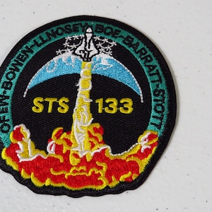 May include: Embroidered patch featuring a space shuttle launching. The patch is circular with a black background and a green border. Text includes "STS 133" and names. The shuttle is white with blue accents, and the flames are red and yellow.